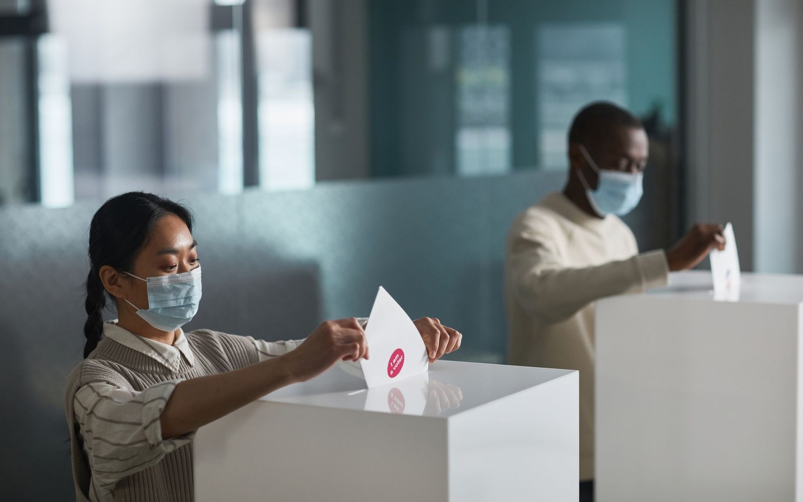 Masked voters casting ballots into separate white ballot boxes indoors