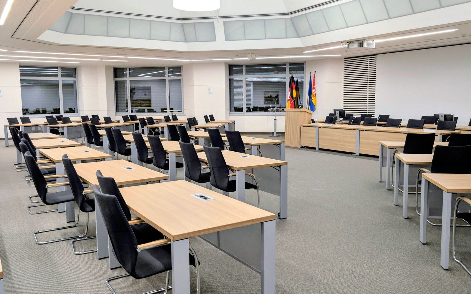 Empty council chamber with rows of desks, chairs and a lectern beside flags