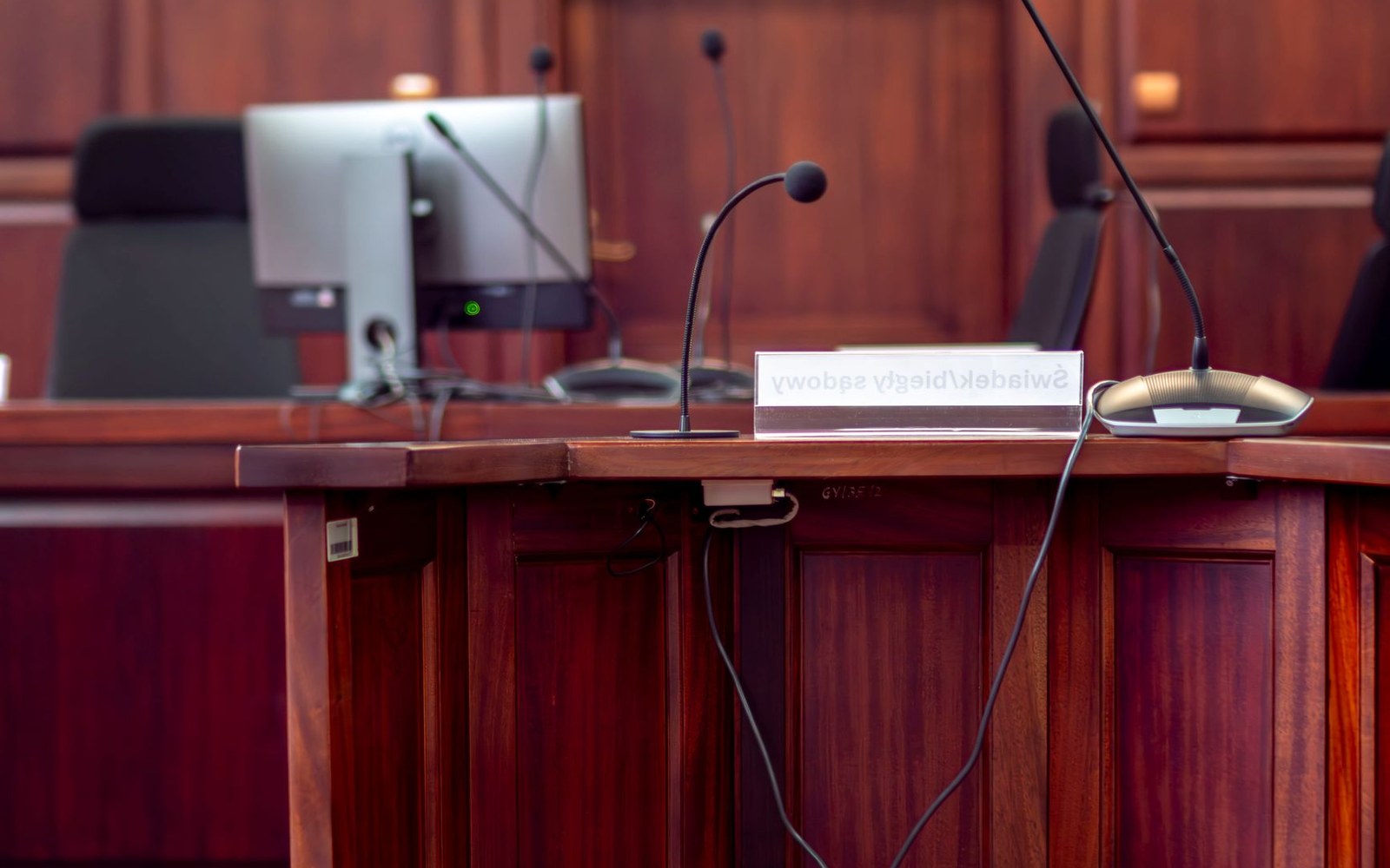 Empty courtroom bench with microphones, a monitor, and a witness nameplate sign