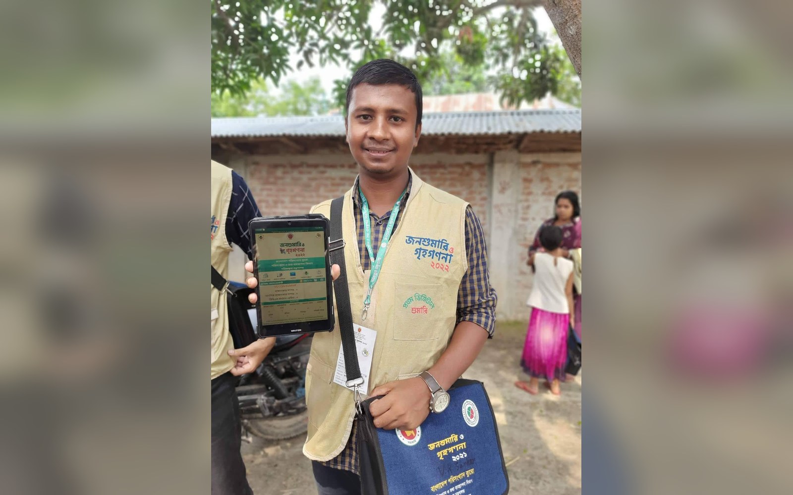 A census enumerator in a branded vest holds up a tablet displaying survey data