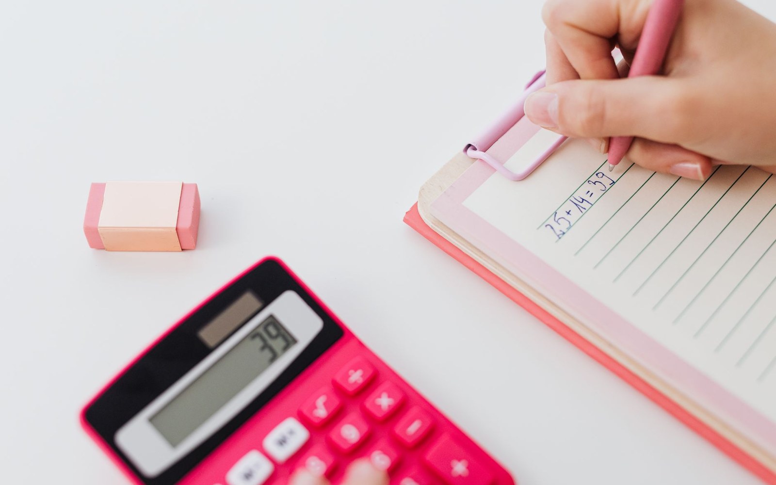 A hand writes an addition sum in a notebook beside a pink calculator and eraser