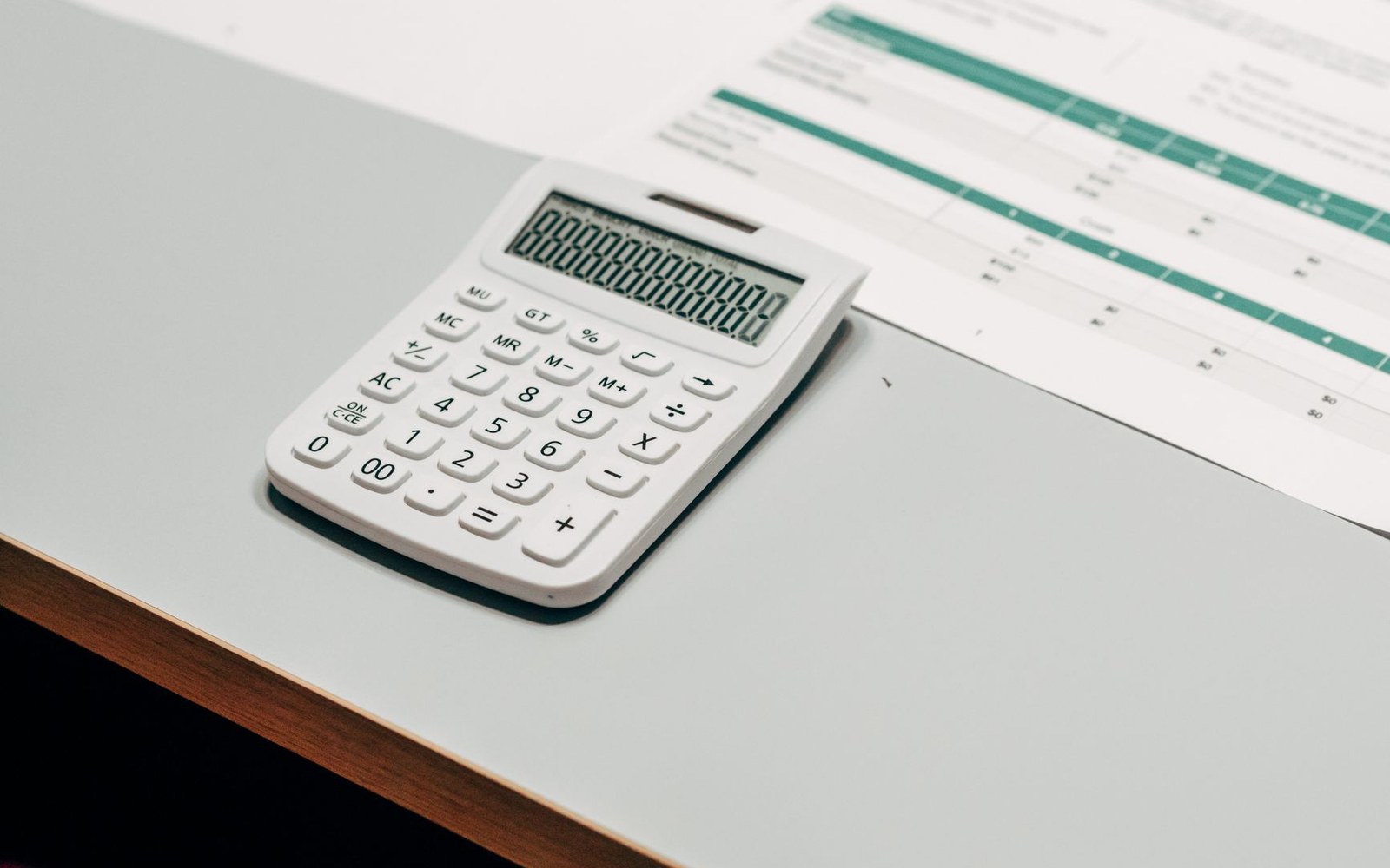A white calculator resting on a desk beside a printed spreadsheet with green highlights