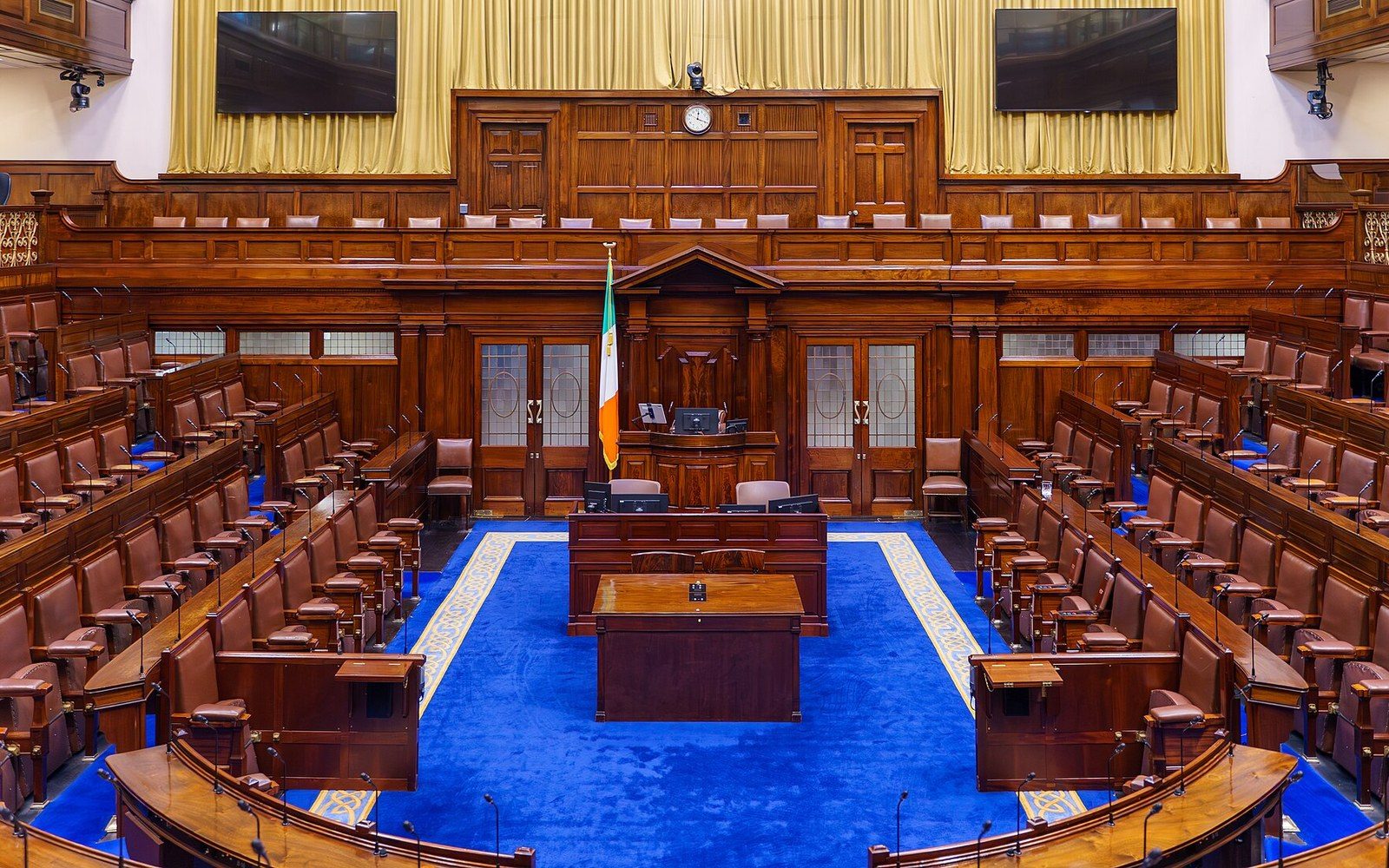 Empty wood-paneled chamber with tiered leather benches around a blue-carpeted floor and Irish flag