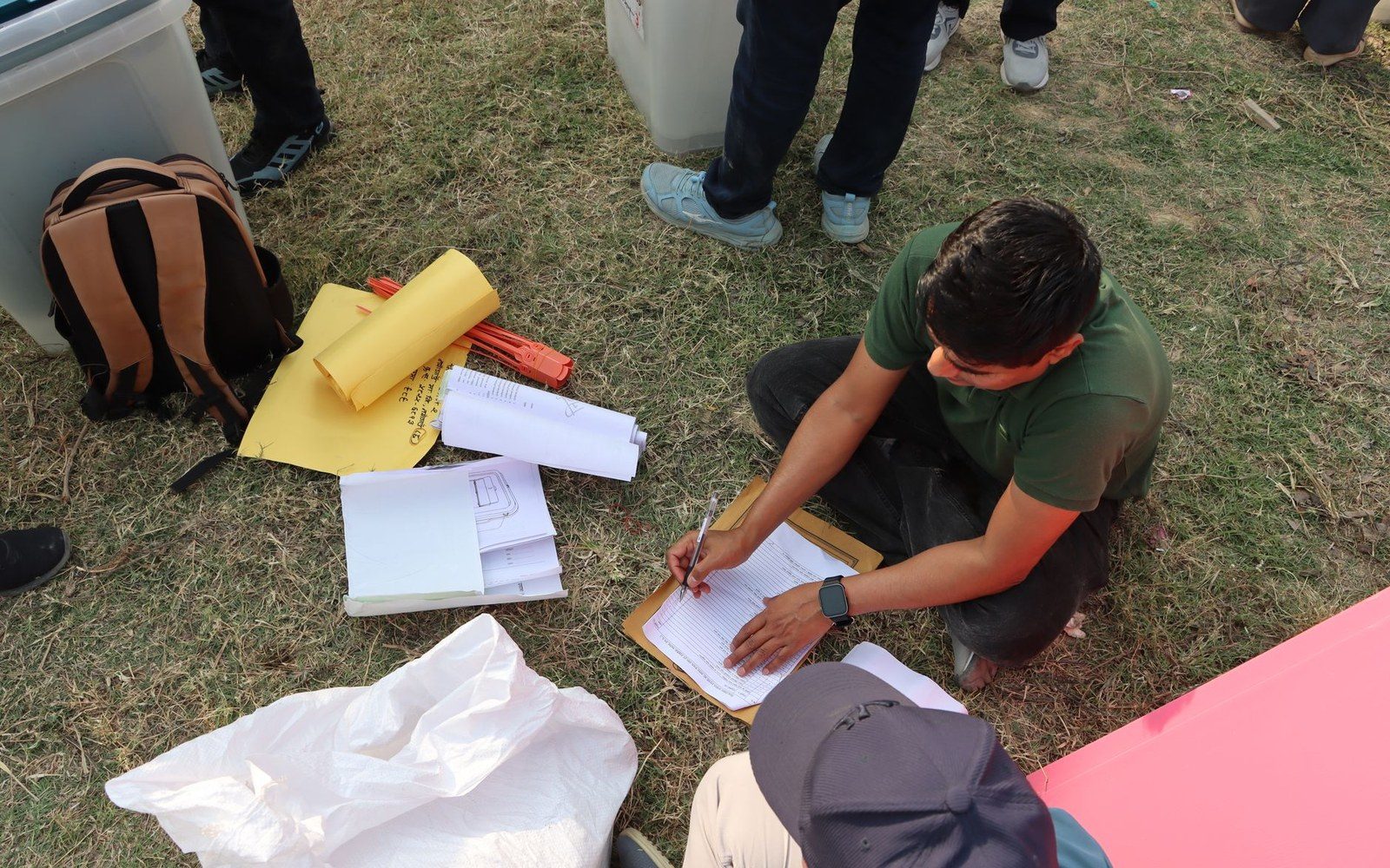 A man kneels on grass filling out paperwork on a clipboard beside scattered documents and bags