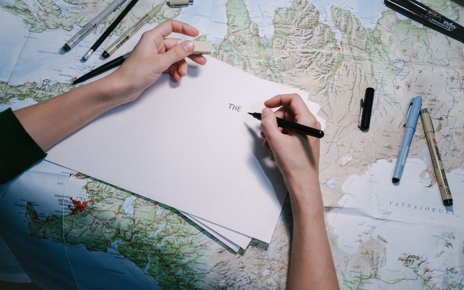 Hands lettering the word "the" on paper atop a map, surrounded by pens